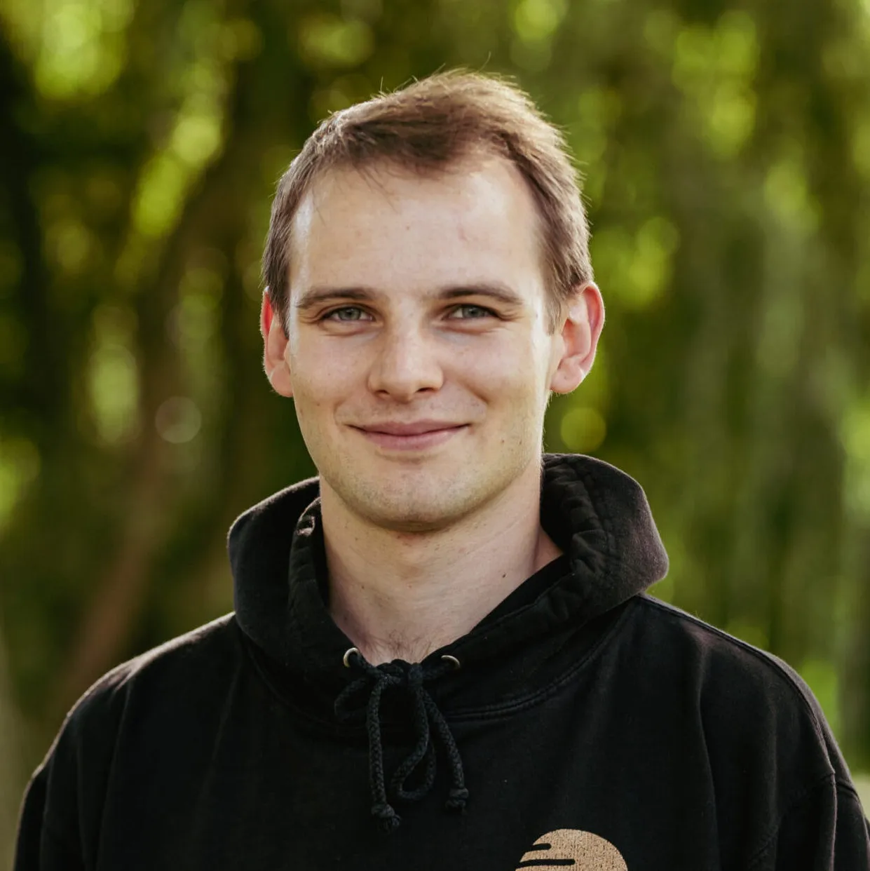Porträt eines jungen Mannes (Julian) mit kurzen Haaren im schwarzen Hoodie, leicht lächelnd, vor unscharfem, grünem Naturhintergrund.